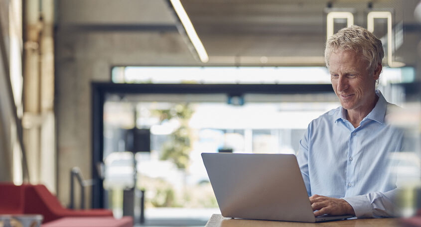 Mature businessman smiling while working on a laptop in a modern office setting with a stylish interior and natural light streaming in from large windows.