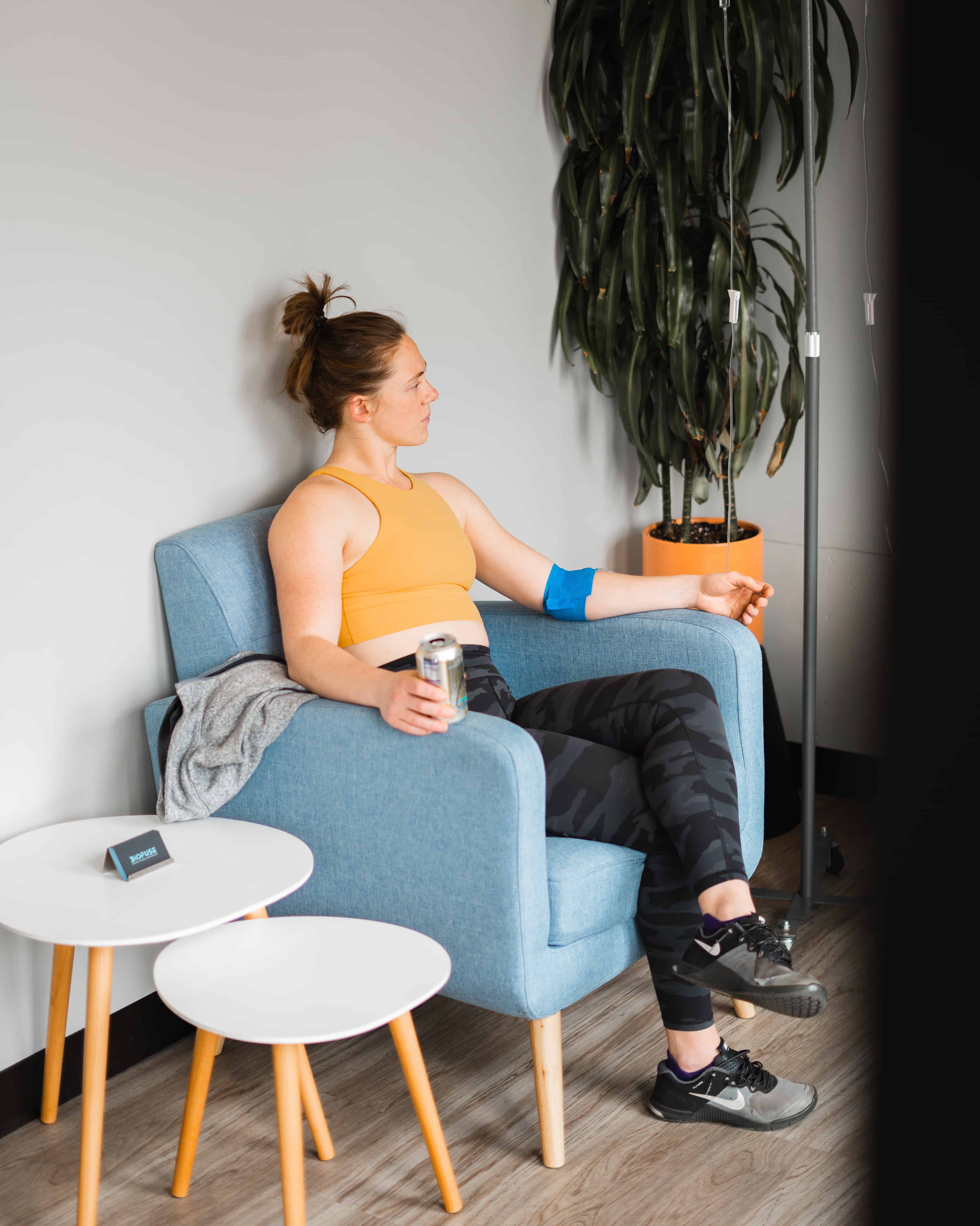 A woman in athletic wear relaxes on a blue armchair with an IV drip in her arm, holding a metal can, beside a large potted plant in a modern indoor setting