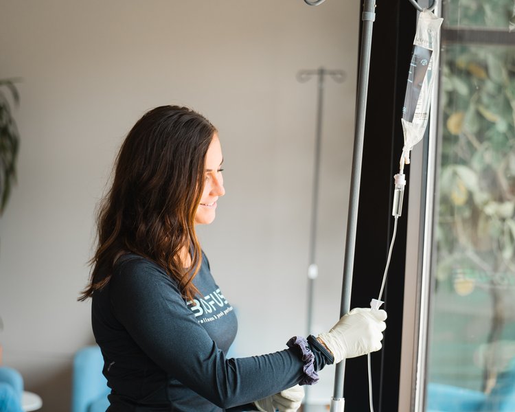 A smiling woman in a casual, long-sleeve t-shirt adjusts an IV stand by a window in a well-lit room