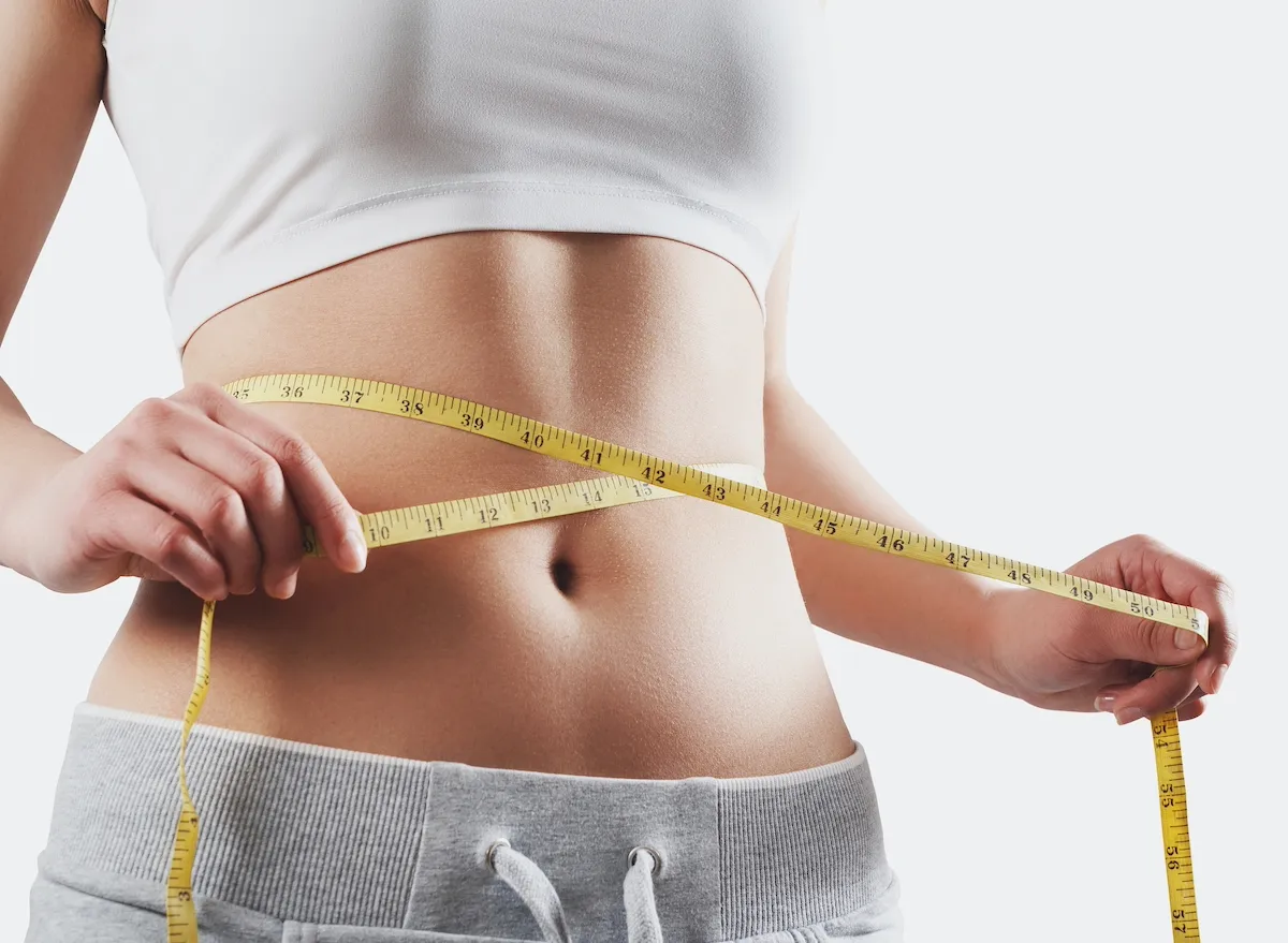 A woman measures her waist with a yellow tape measure, highlighting a focus on physical fitness and body measurement